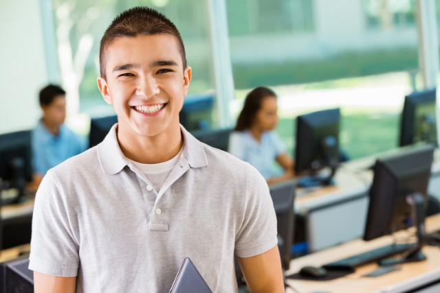 A male student stands in his classroom and smiles at the camera.