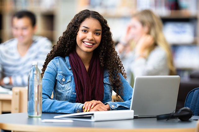 A young African American female student sits and works on her laptop in the library of her school.