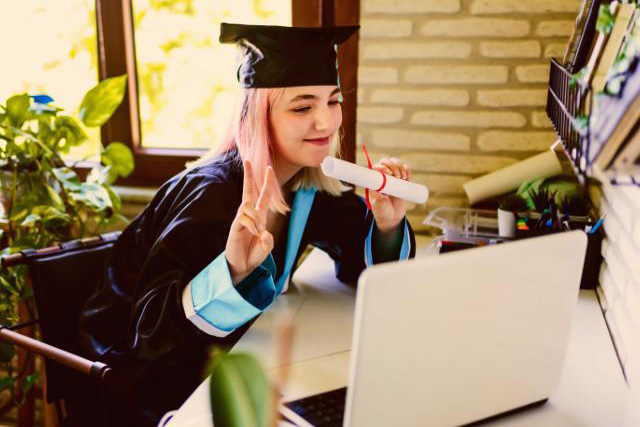 A girl student with diploma is sitting in front of her laptop smiling and does the victory sign with her fingers.