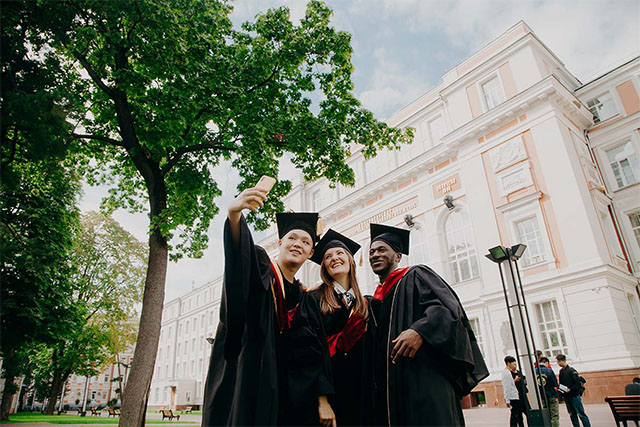 Three graduates, wearing their cap, is taking a selfie, in front of school building.