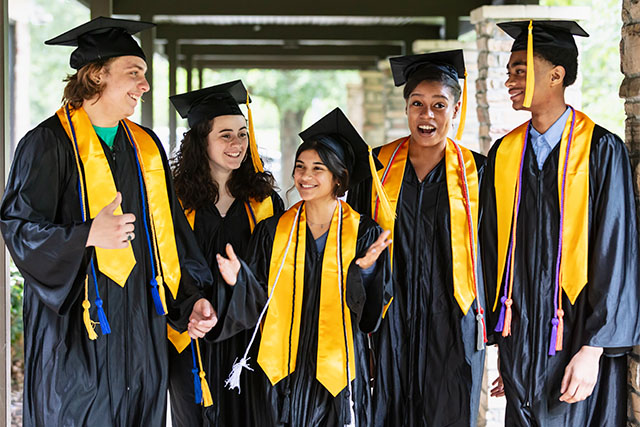 A group of five multiracial high school graduates standing together outside the school, wearing their caps and gowns, smiling.