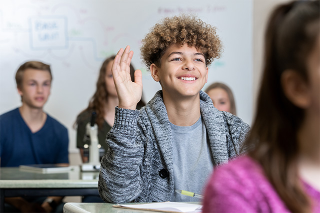 Male high school student smiles while raising hand in class.