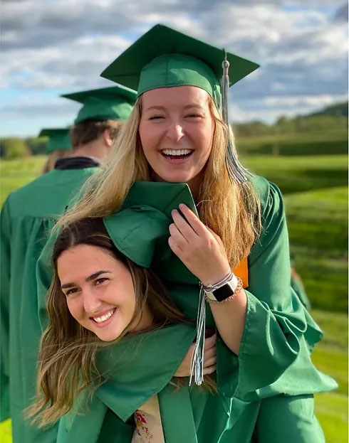Two Academy female students laughing at graduation and is wearing green cap and gown.